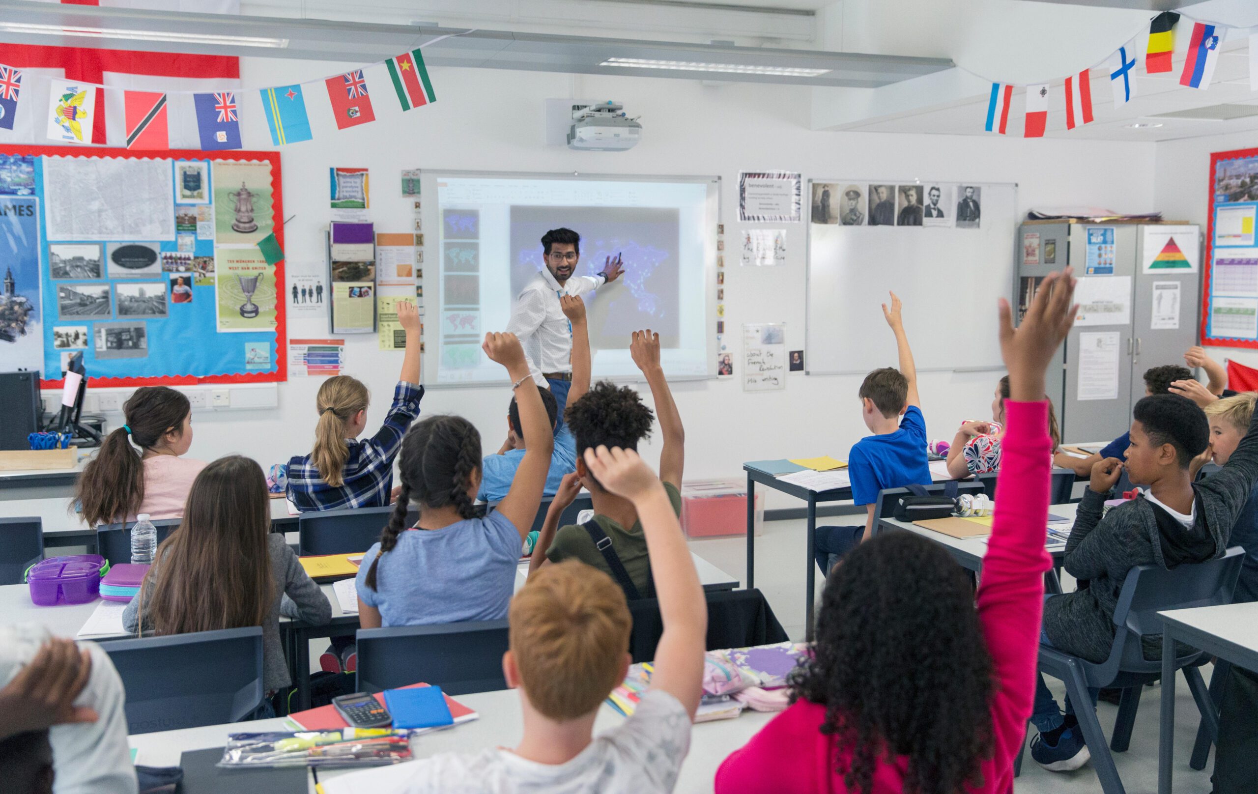 Male teacher delivering a lesson in a classroom with students actively participating by raising their hands.